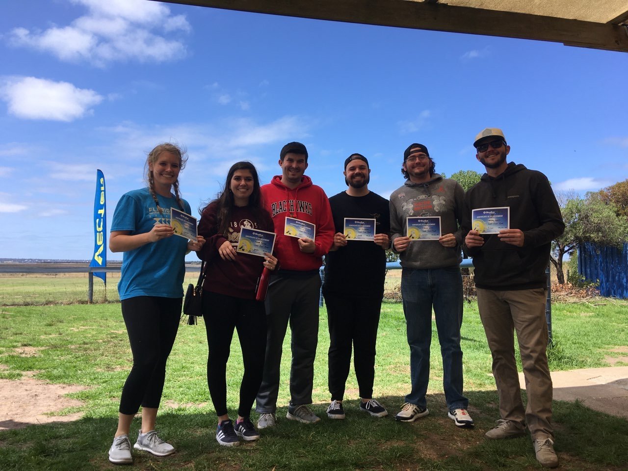 Six friends holding up their skydiving certificates on the grass, a skydive flag behind them.