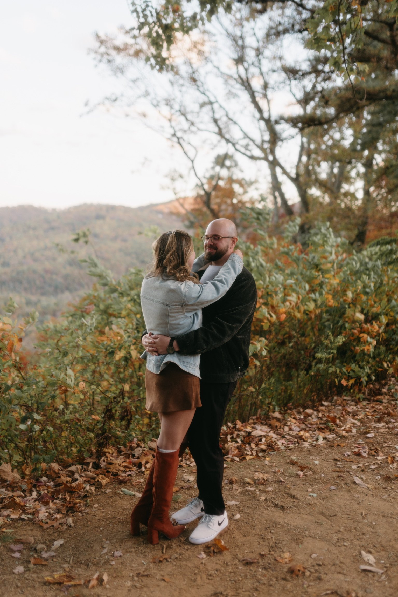 Ian holding Nicole close at the edge of the overlook, autumn leaves at their feet.