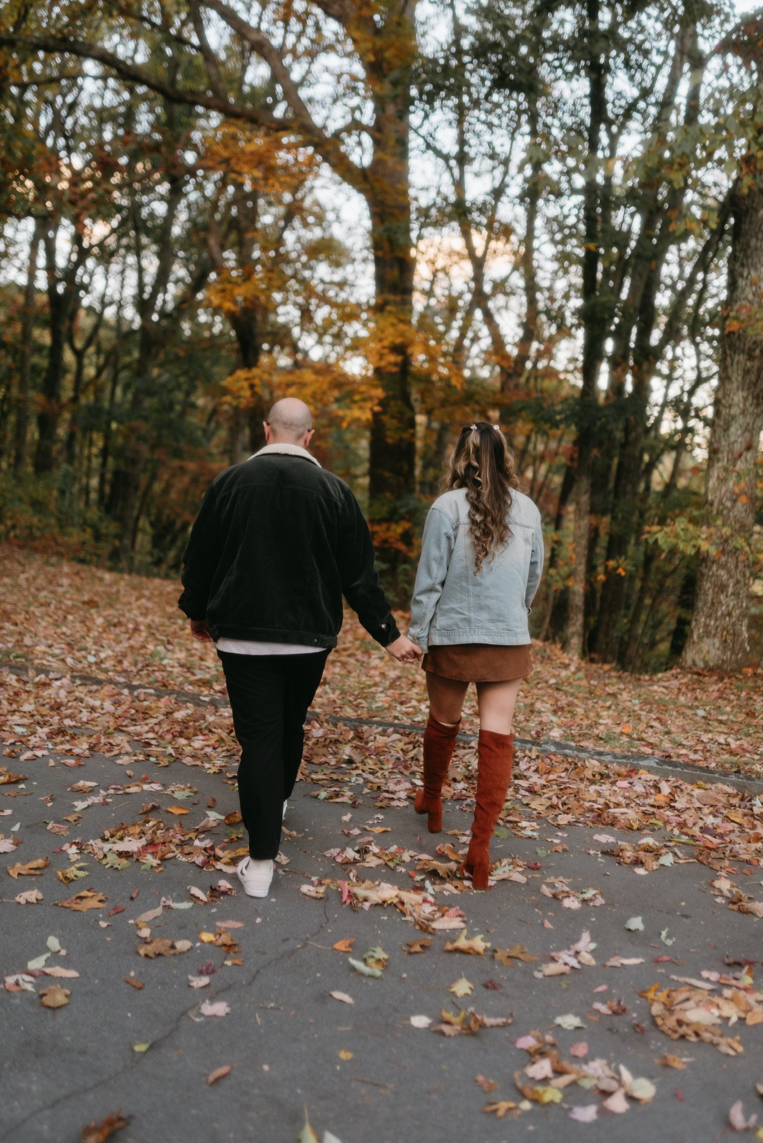 From behind: Nicole and Ian walking hand in hand along a leaf-strewn road through the woods.