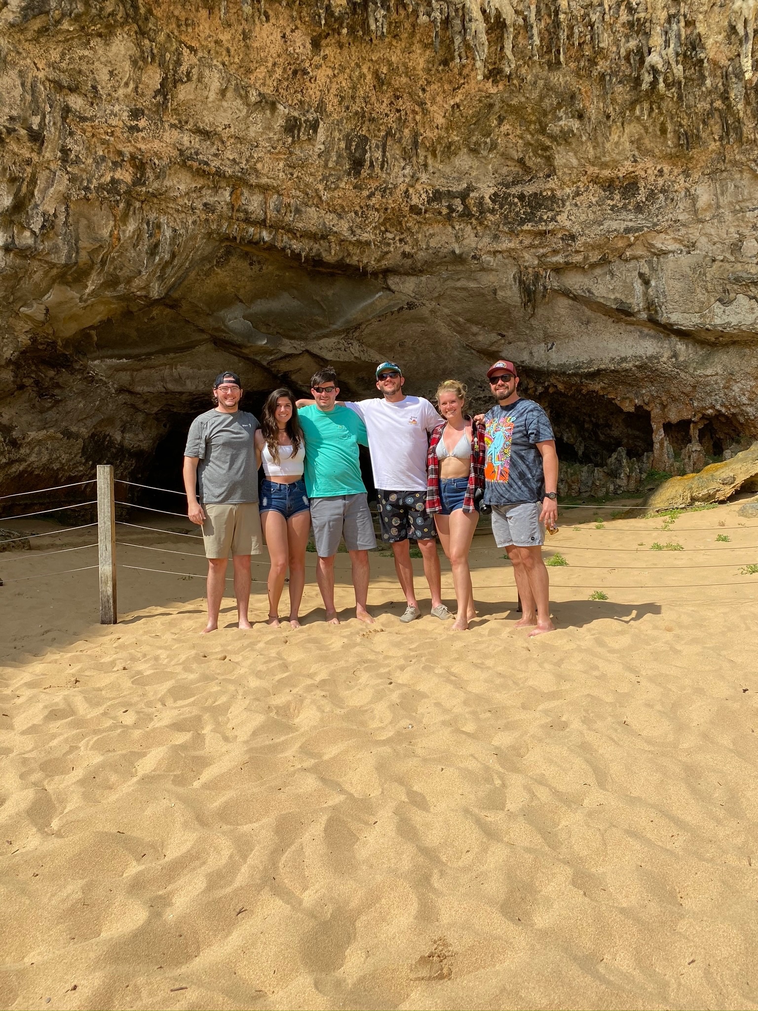 Six friends lined up on the sand beneath an overhanging sandstone cliff.