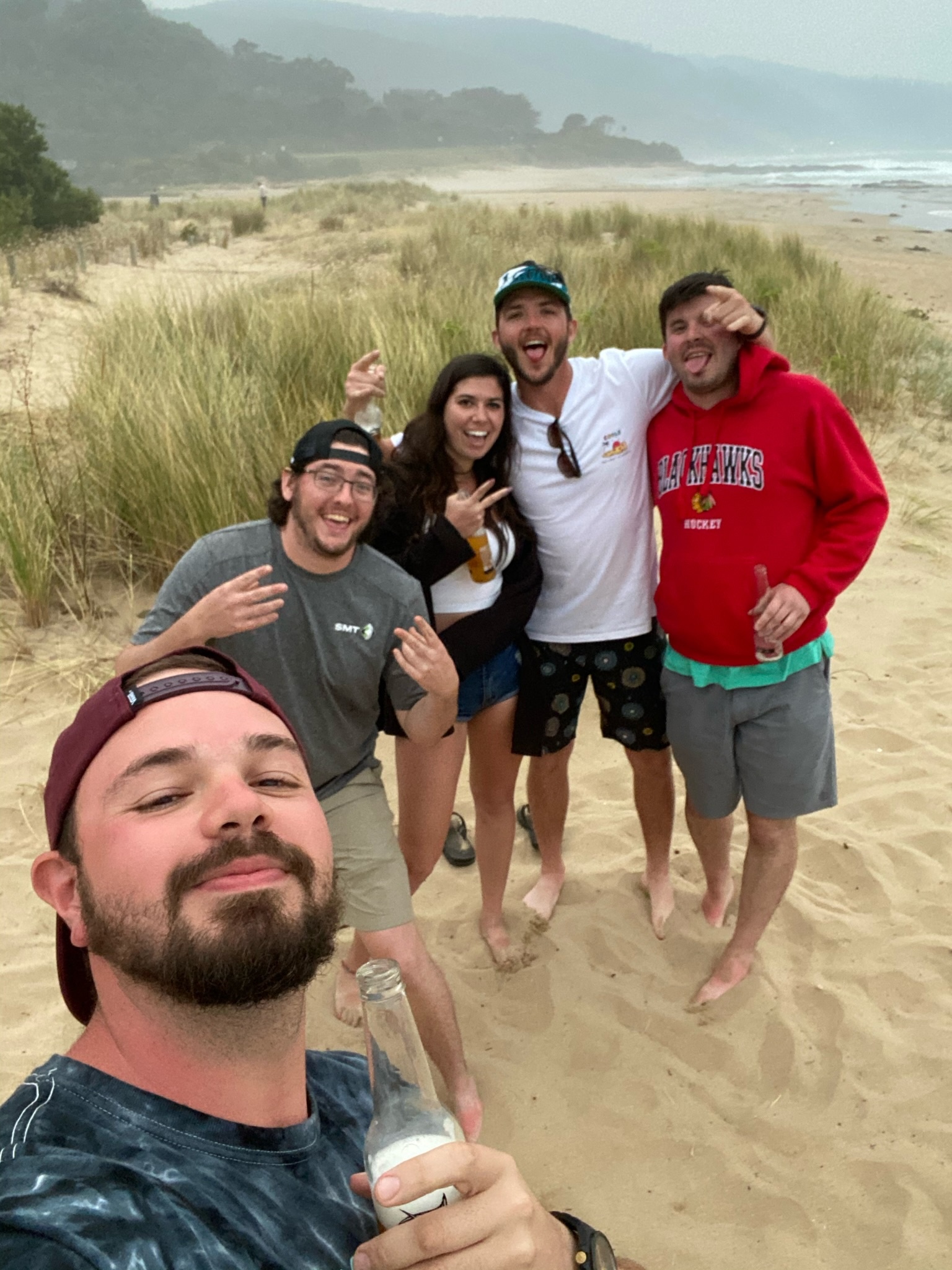 A laughing selfie of friends in the dunes, drinks in hand, a misty beach behind them.