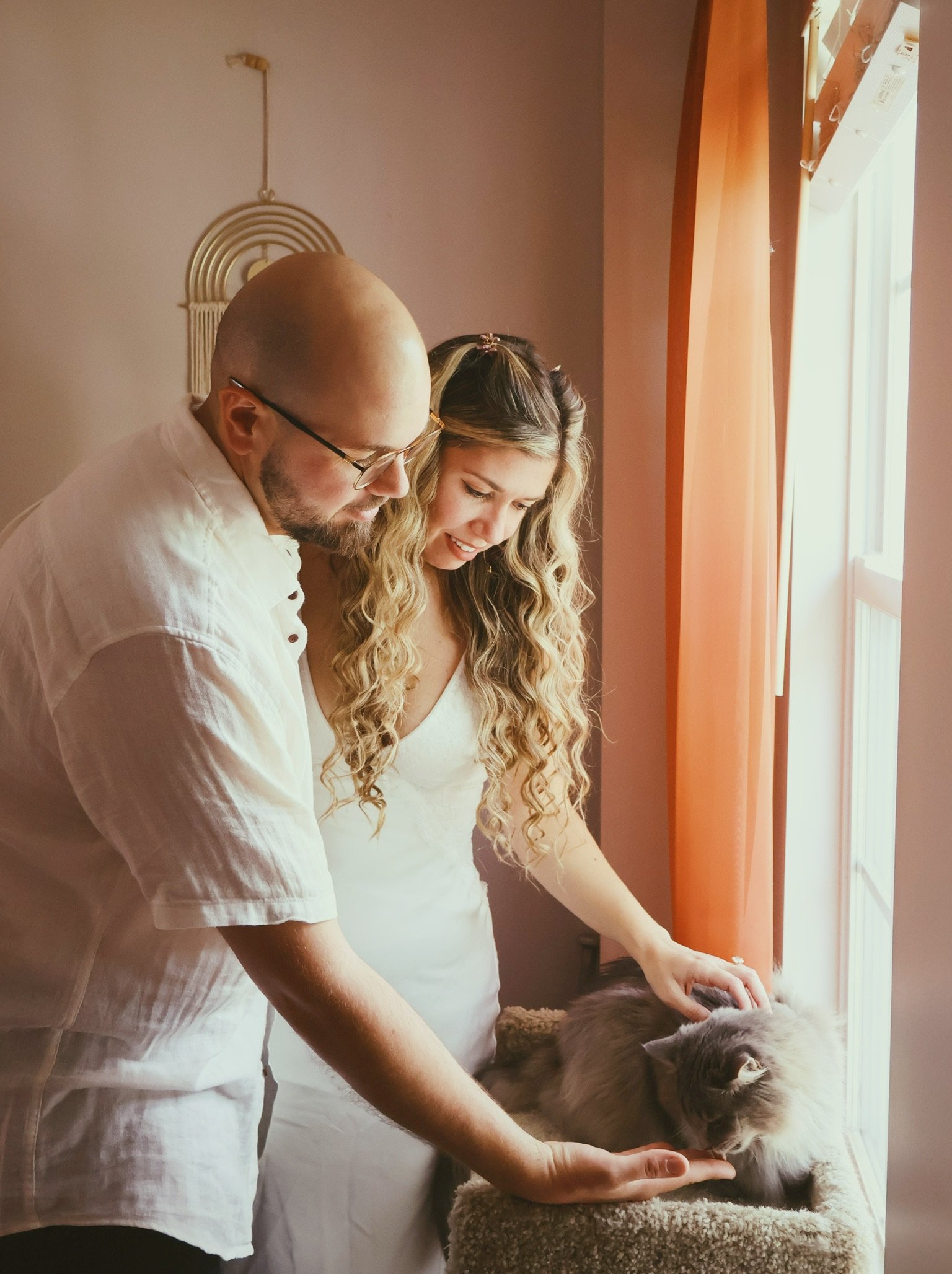 Ian and Nicole in white by the window, reaching down to stroke a cat on its perch.