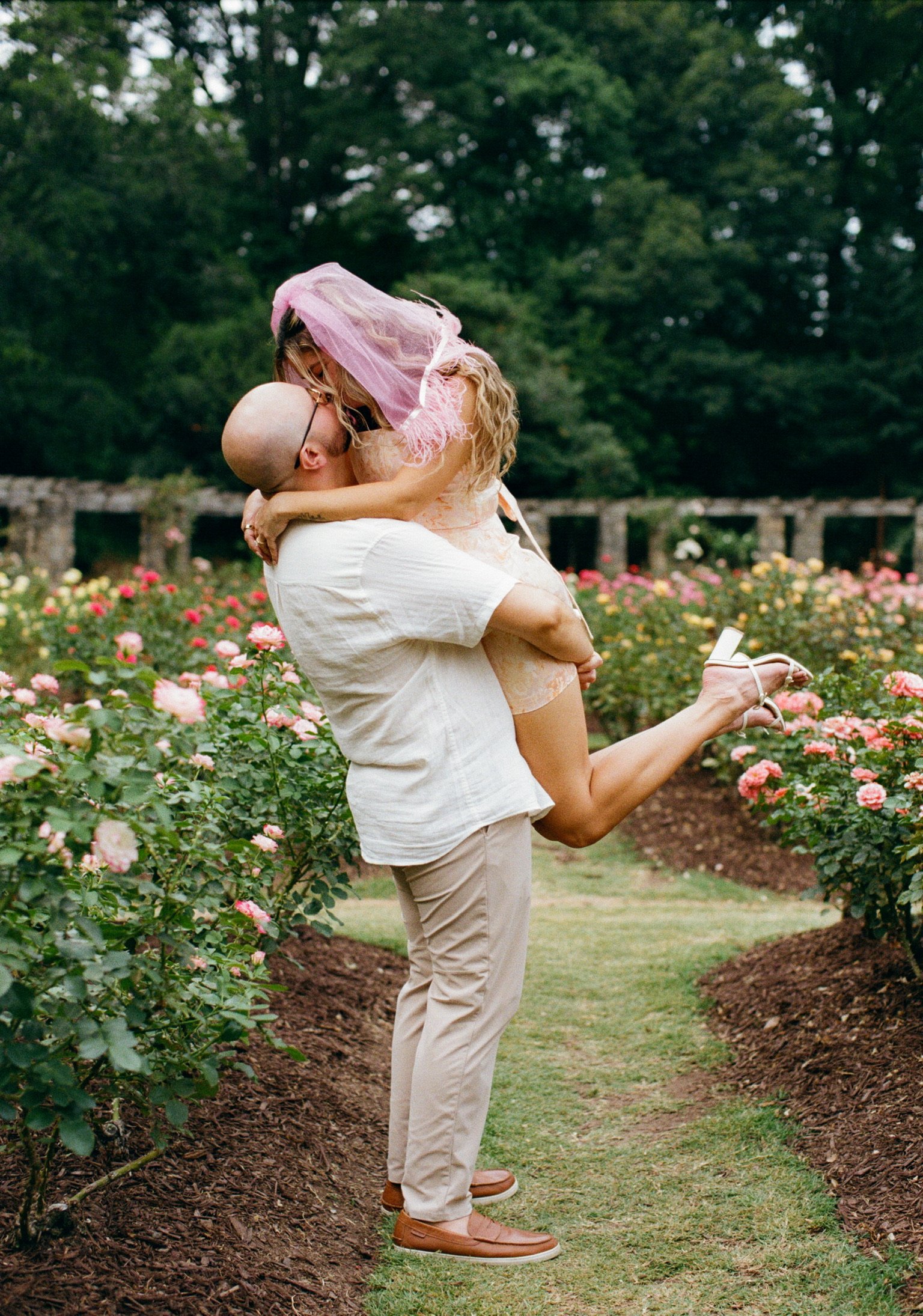Ian lifting Nicole off her feet in the middle of a grass path between the rose beds.