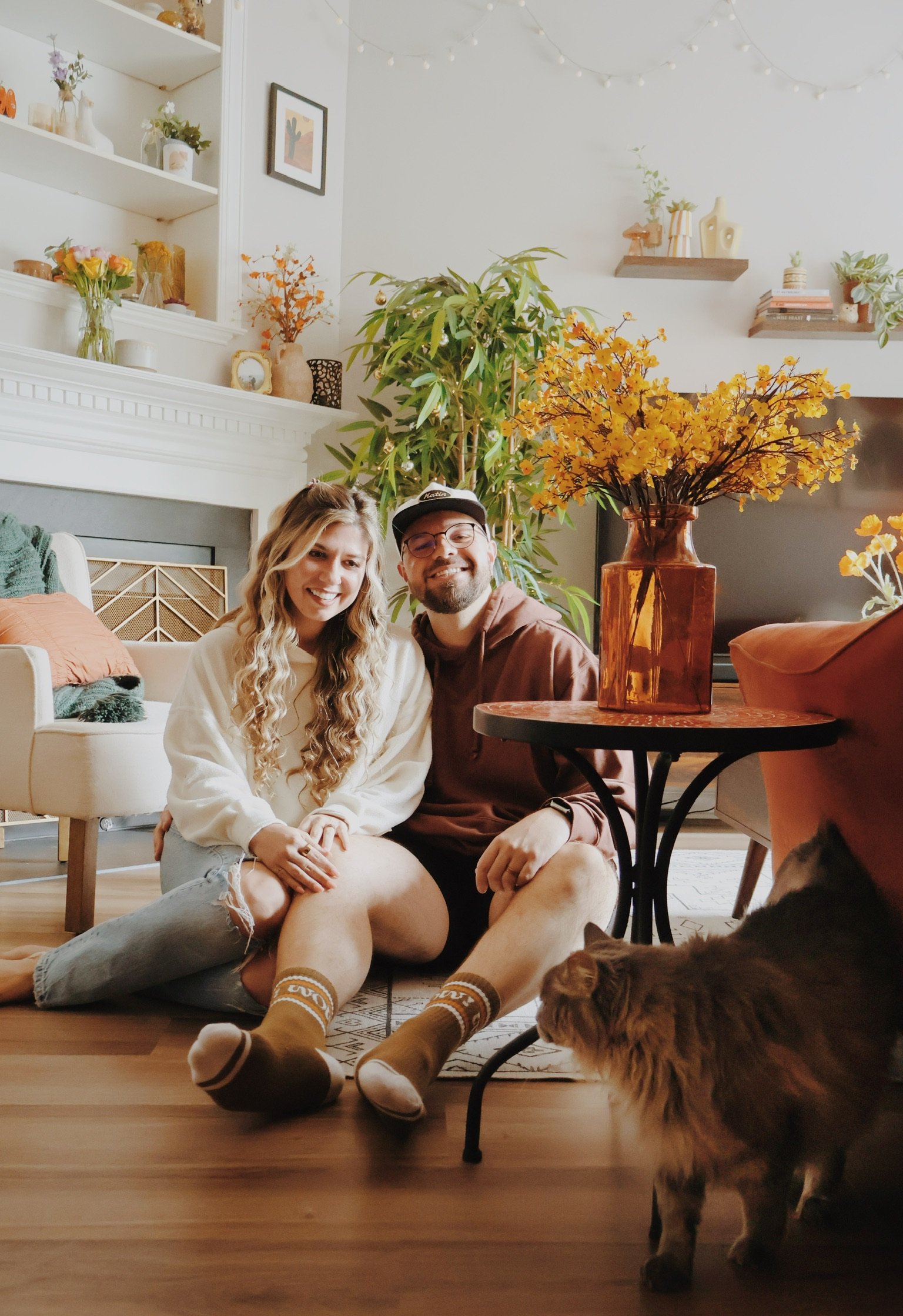 Nicole and Ian sitting on the living-room floor in matching socks while one of their cats wanders past, yellow flowers on the table behind.