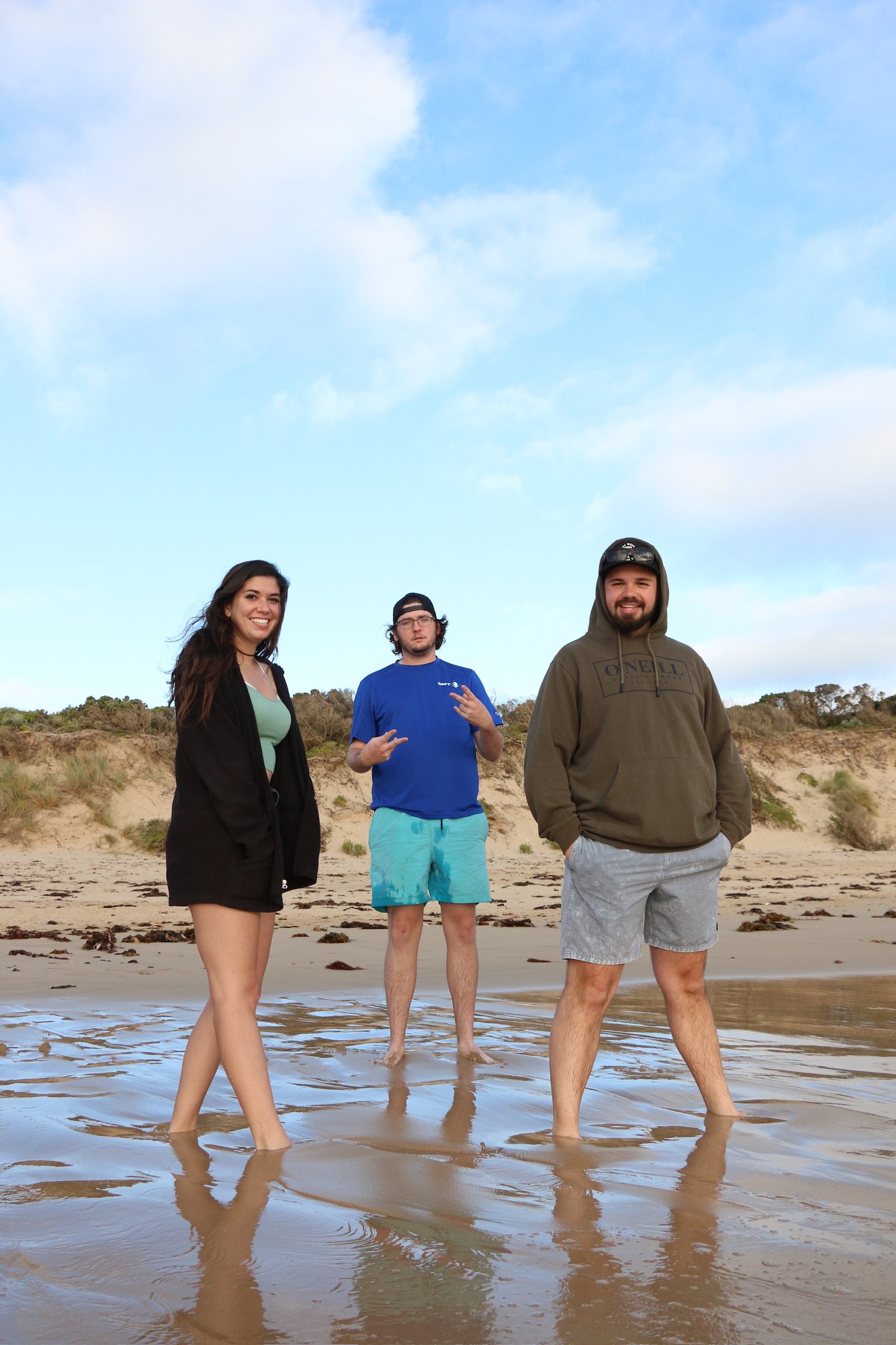 Three friends standing barefoot on wet sand under a big blue sky, their reflections in the water.
