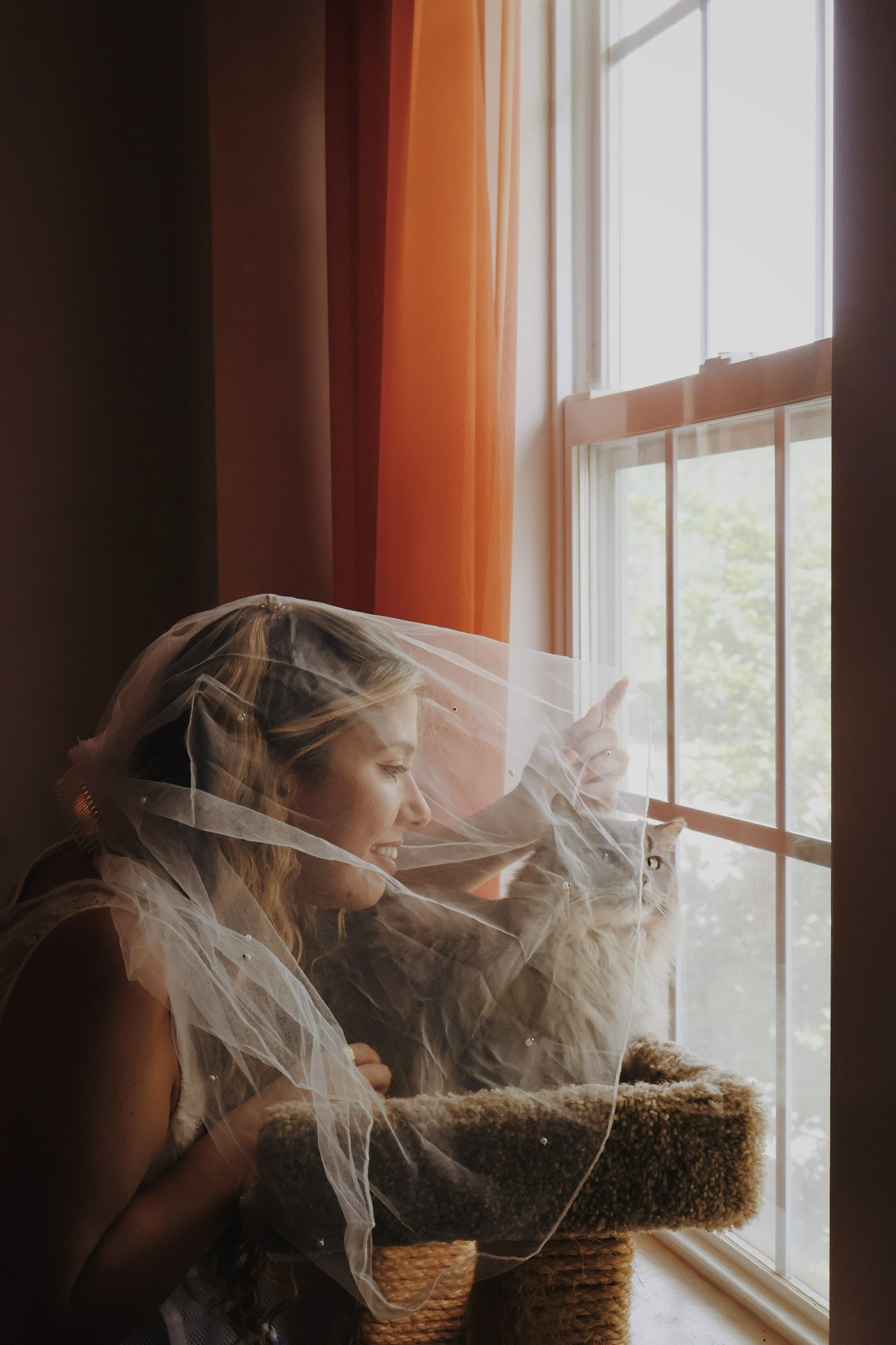 Nicole in a bridal veil at the window, leaning in to a cat on its window perch.