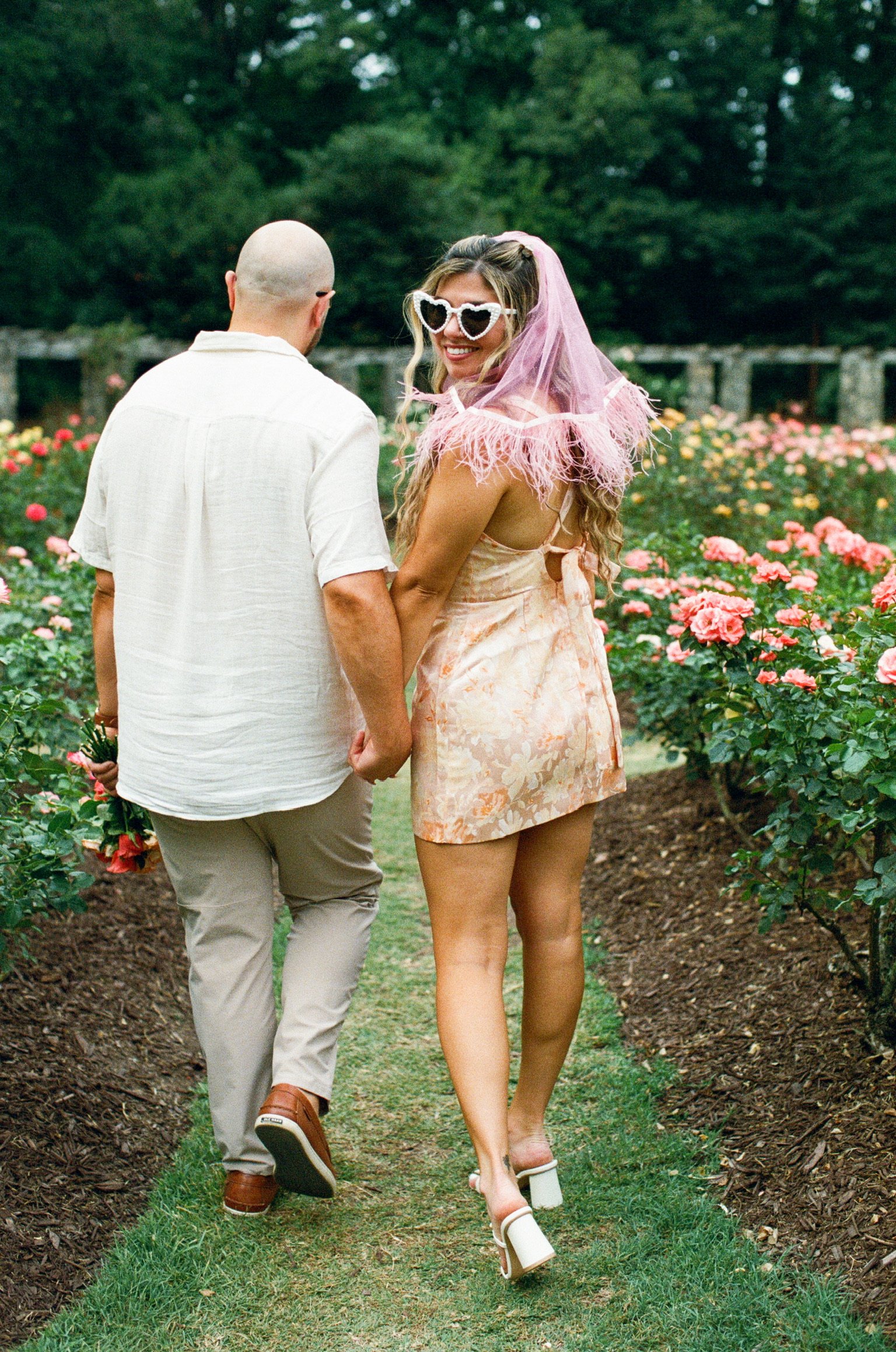 From behind: Nicole in her pink veil and Ian walking hand in hand down a grass path between the roses.