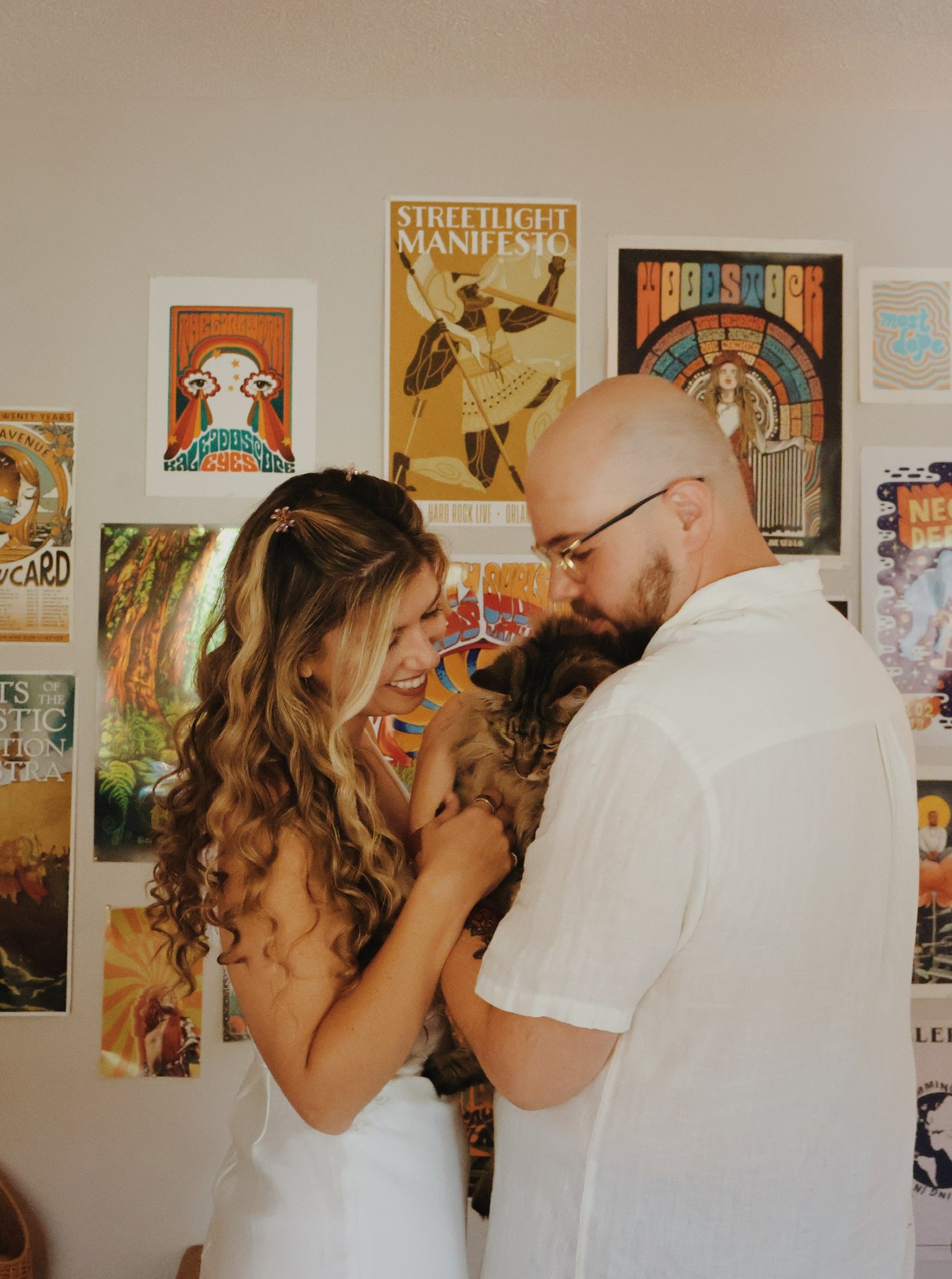 Nicole and Ian in white, cuddling a cat between them in front of the gig posters.