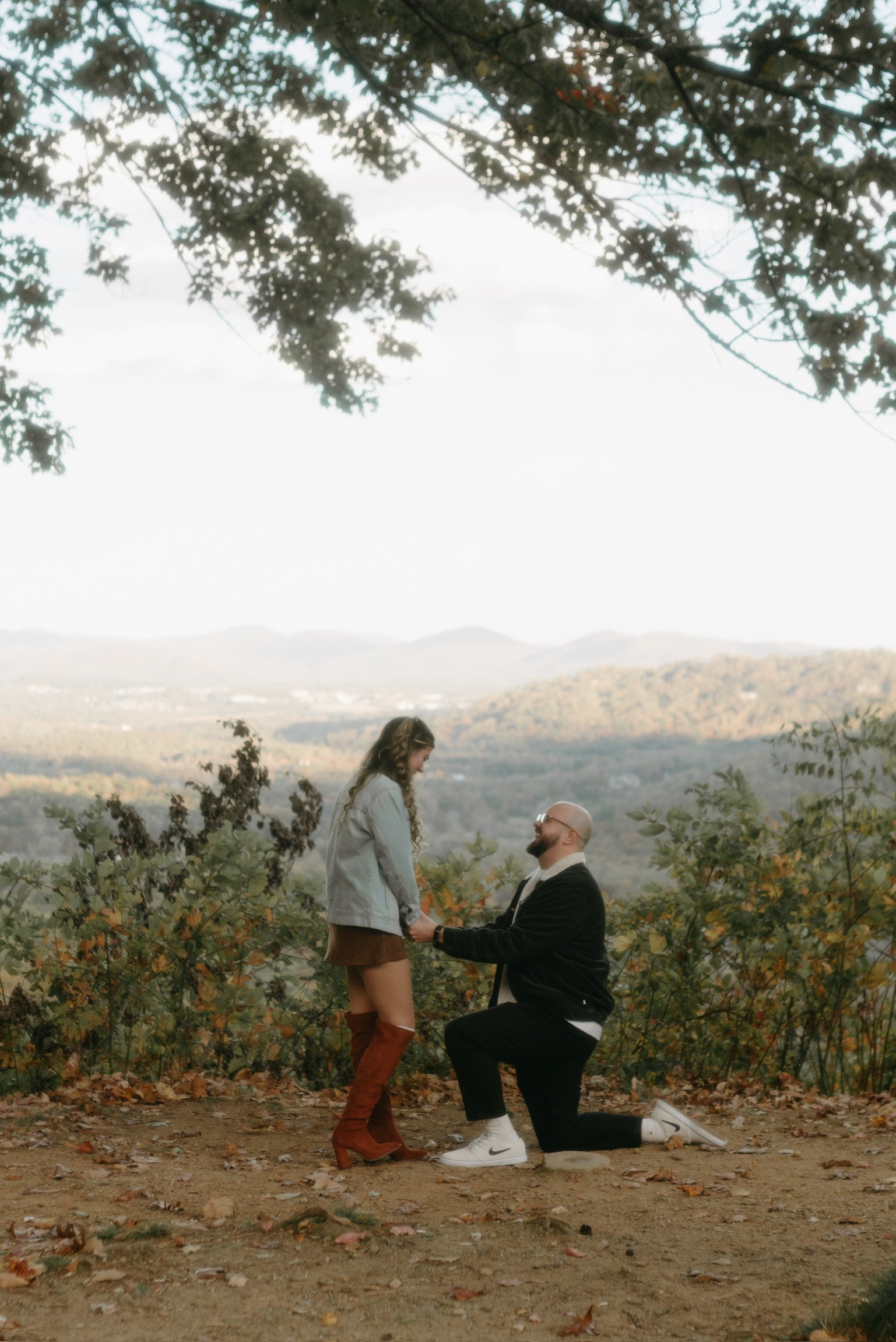 Ian on one knee at the edge of the overlook, Nicole in a denim jacket and rust boots, the ridges fading behind them.