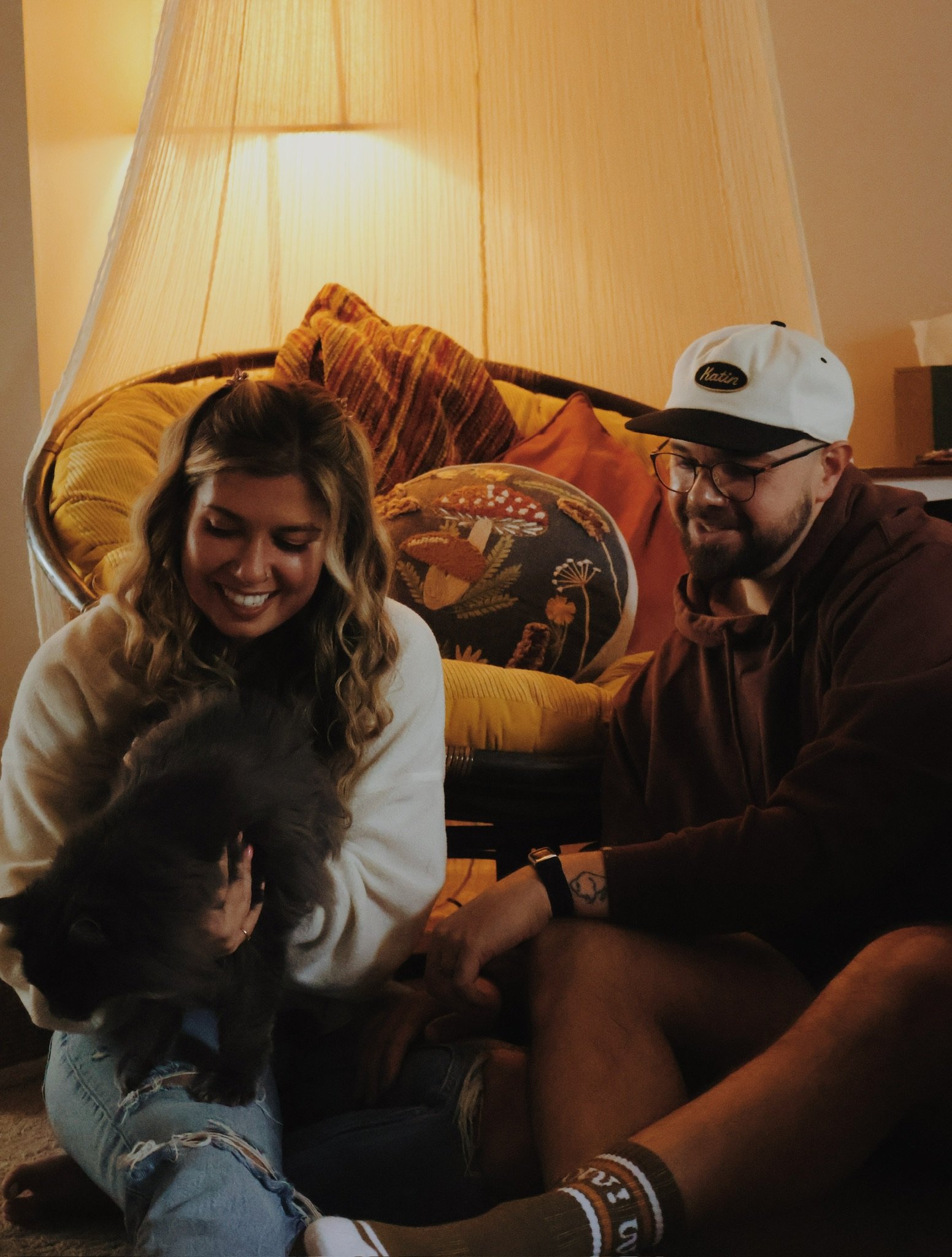Nicole and Ian laughing on the floor by the papasan chair, the gray cat squirming in Nicole’s arms.