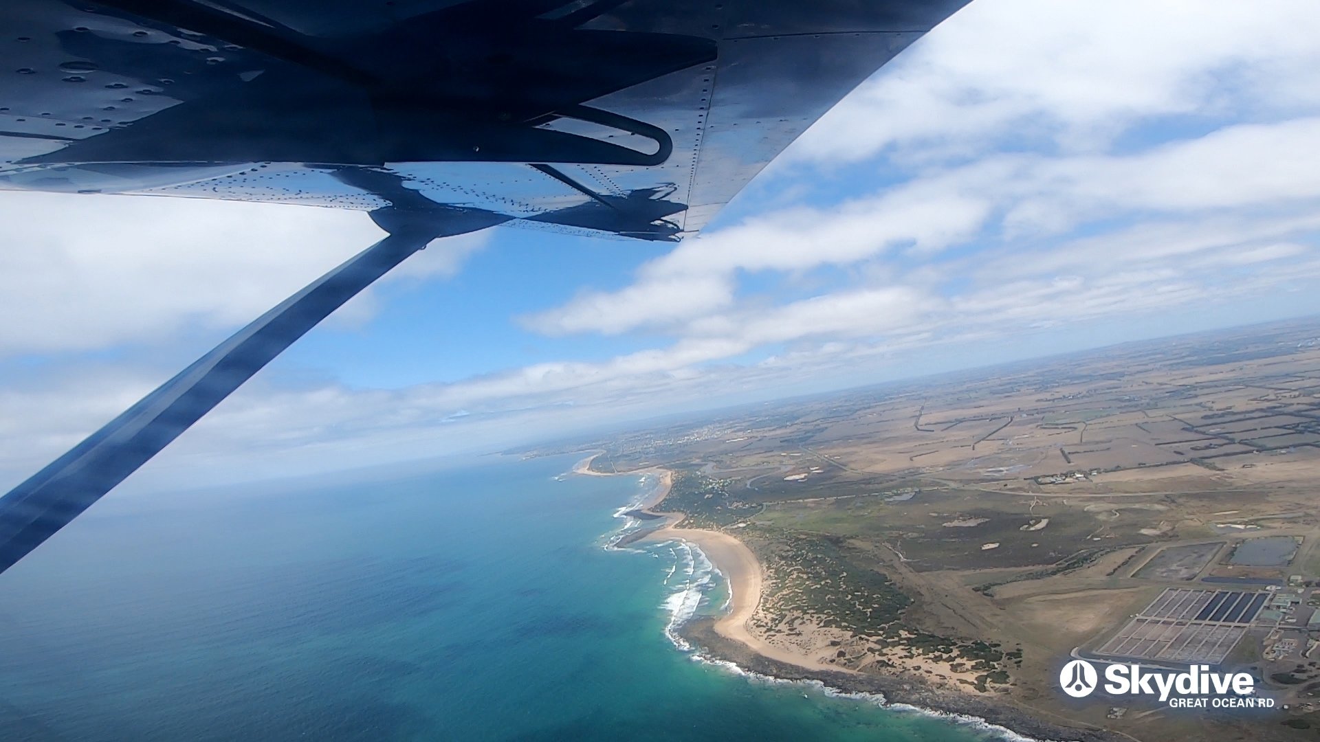 The coastline from the open door of a skydiving plane, turquoise water and a curve of beach far below.