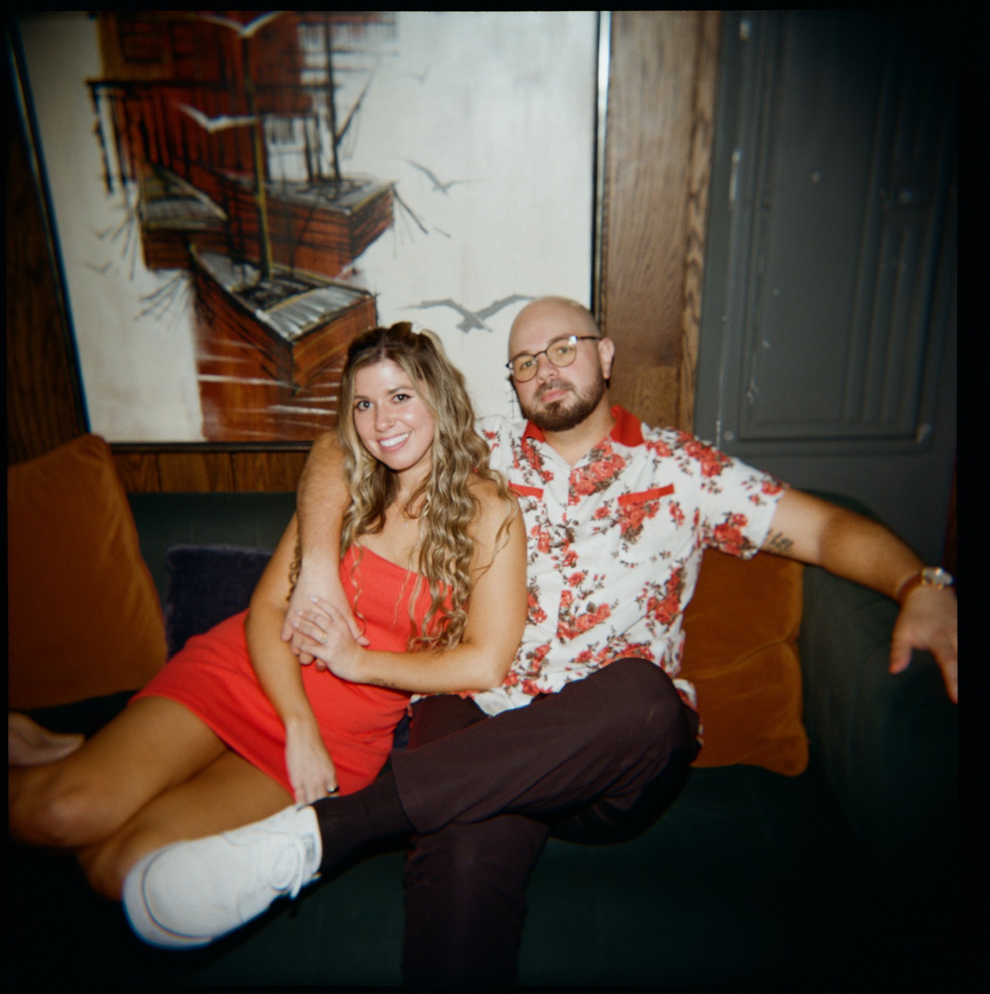 Nicole and Ian sprawled on a velvet banquette under a seagull painting, her arm around his neck.