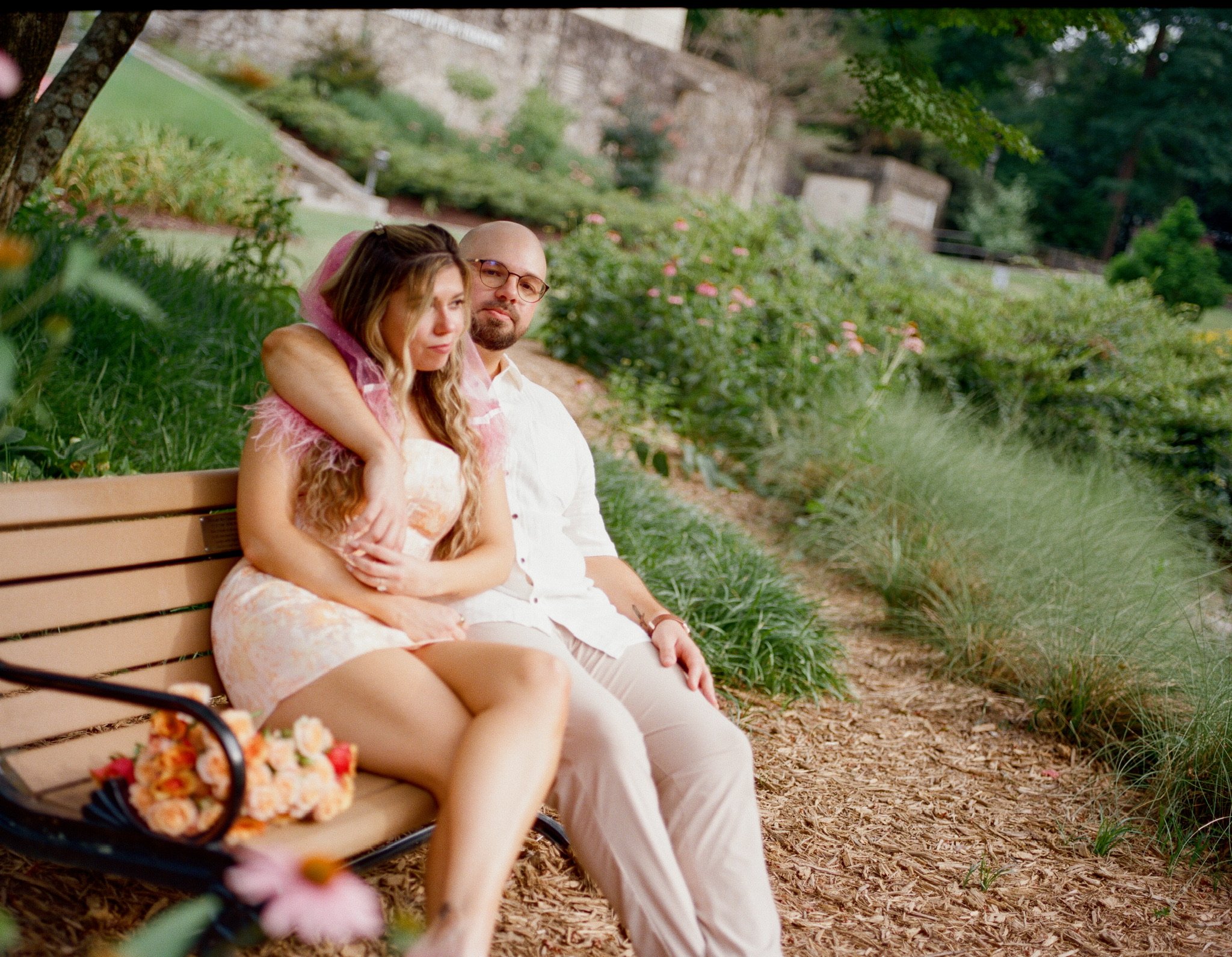 Nicole and Ian sitting on a park bench by the garden path, her legs across his, the bouquet beside them.
