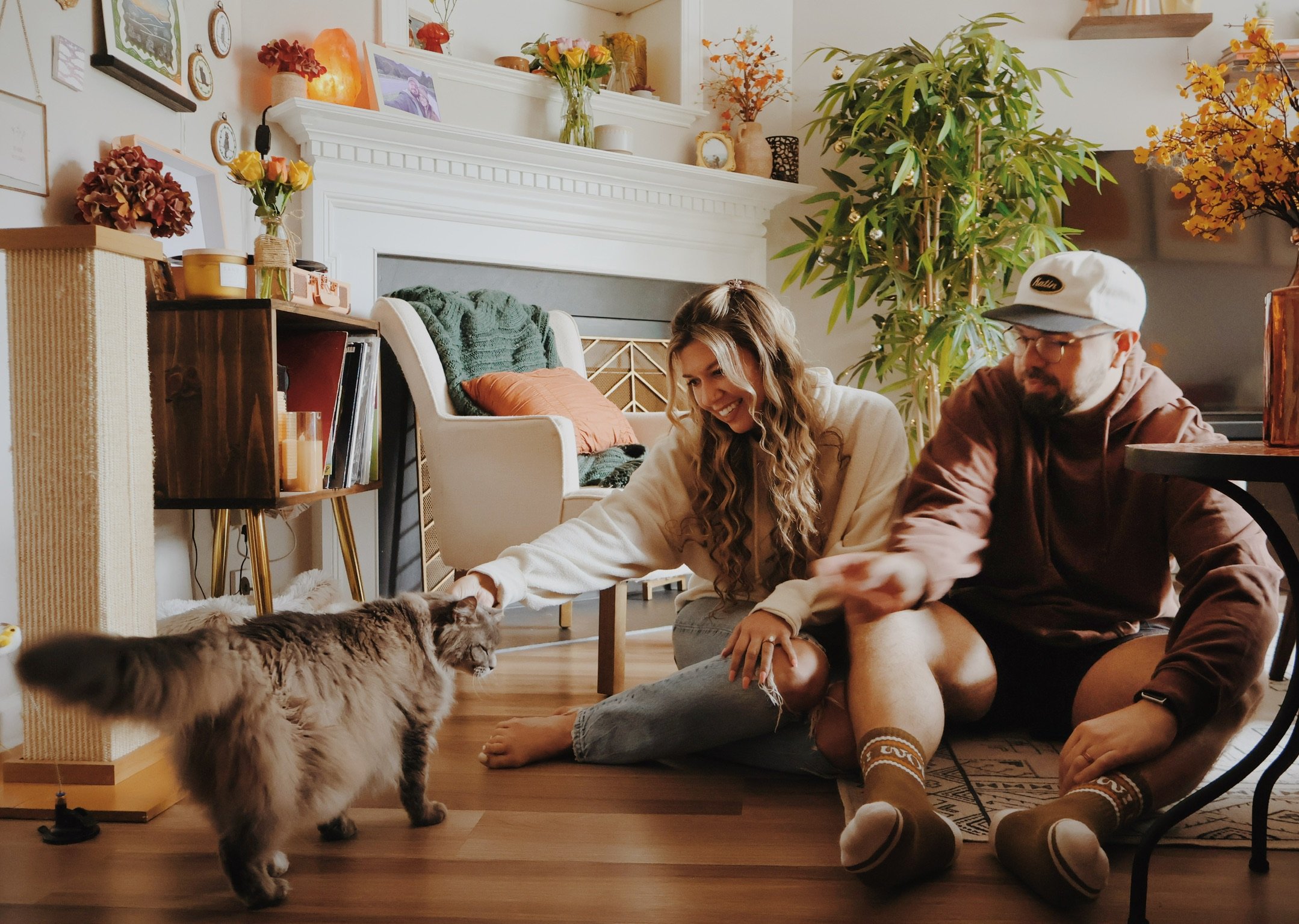 Nicole and Ian sitting on the floor, both reaching for a long-haired cat as it strolls across the room.