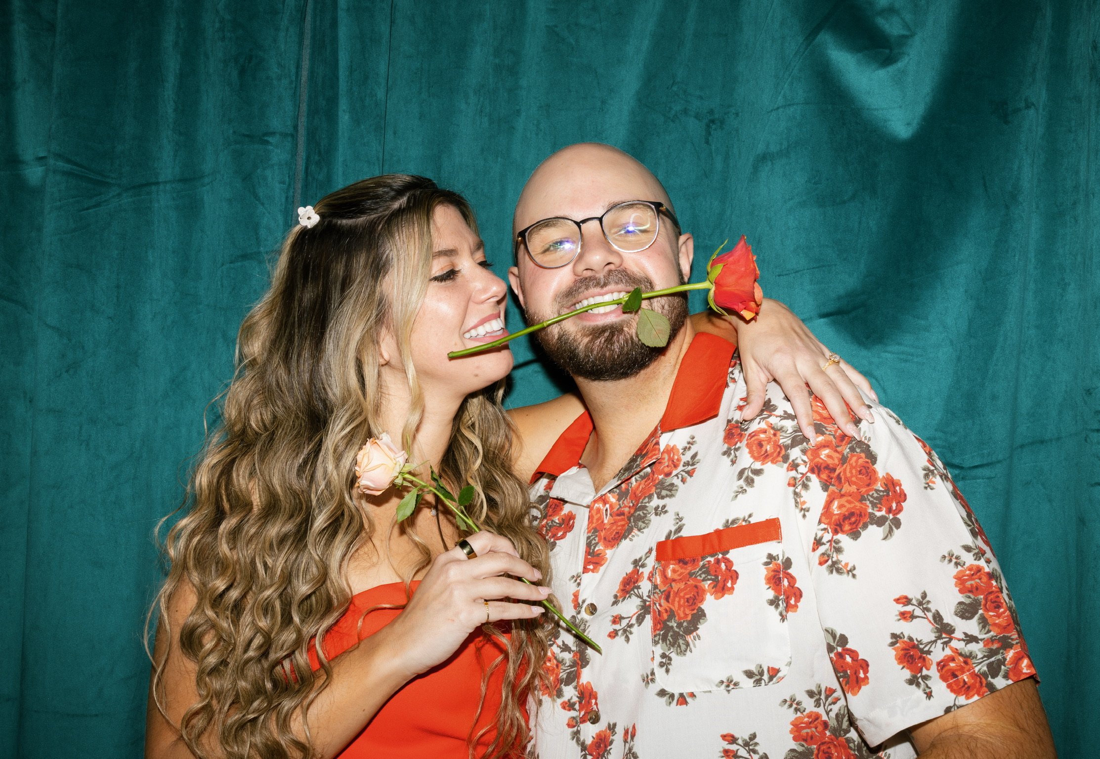 Nicole and Ian against a teal velvet curtain, a red rose clenched between his teeth, both laughing.