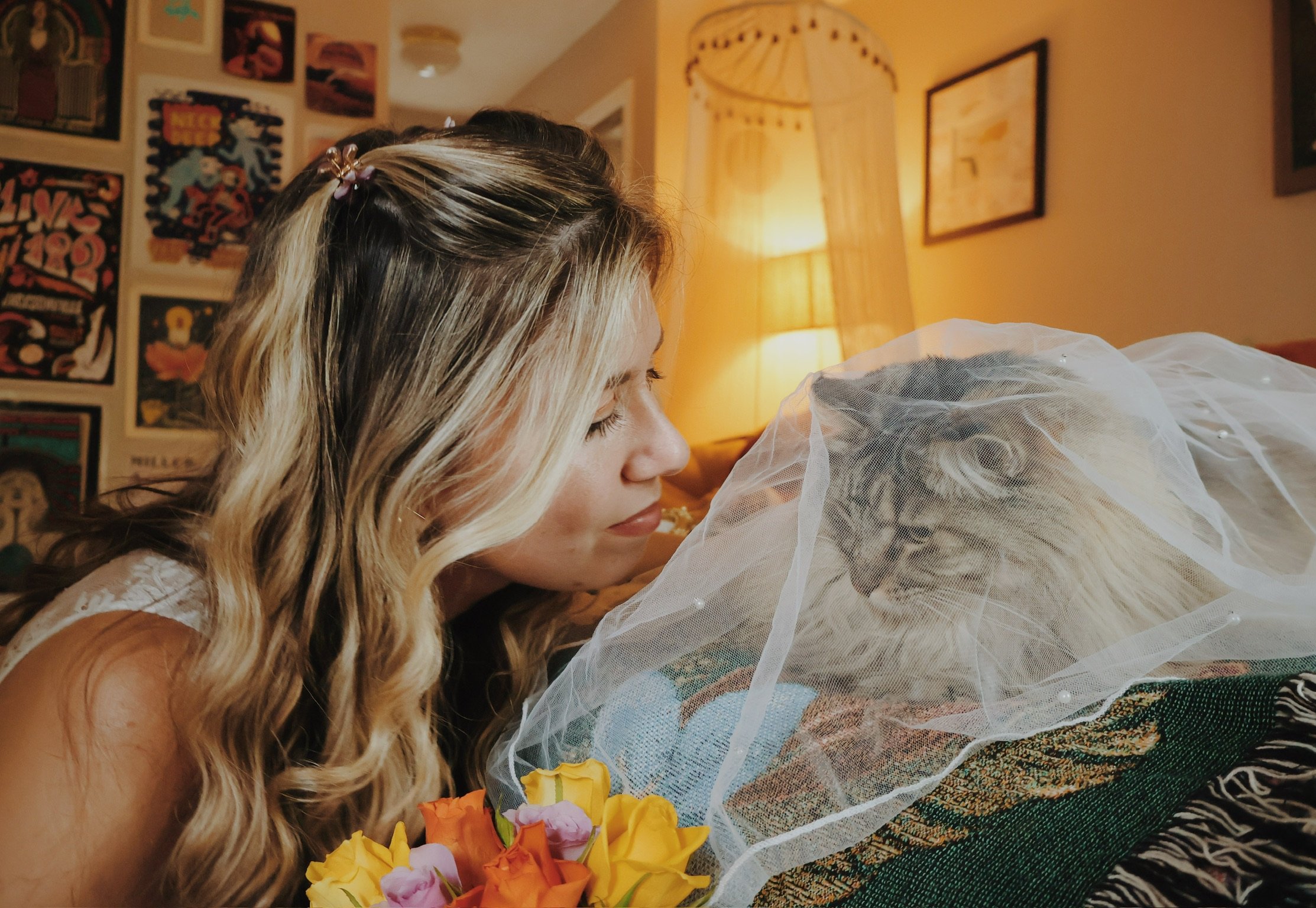Nicole resting her chin beside the veiled cat on the bed, flowers in the foreground.