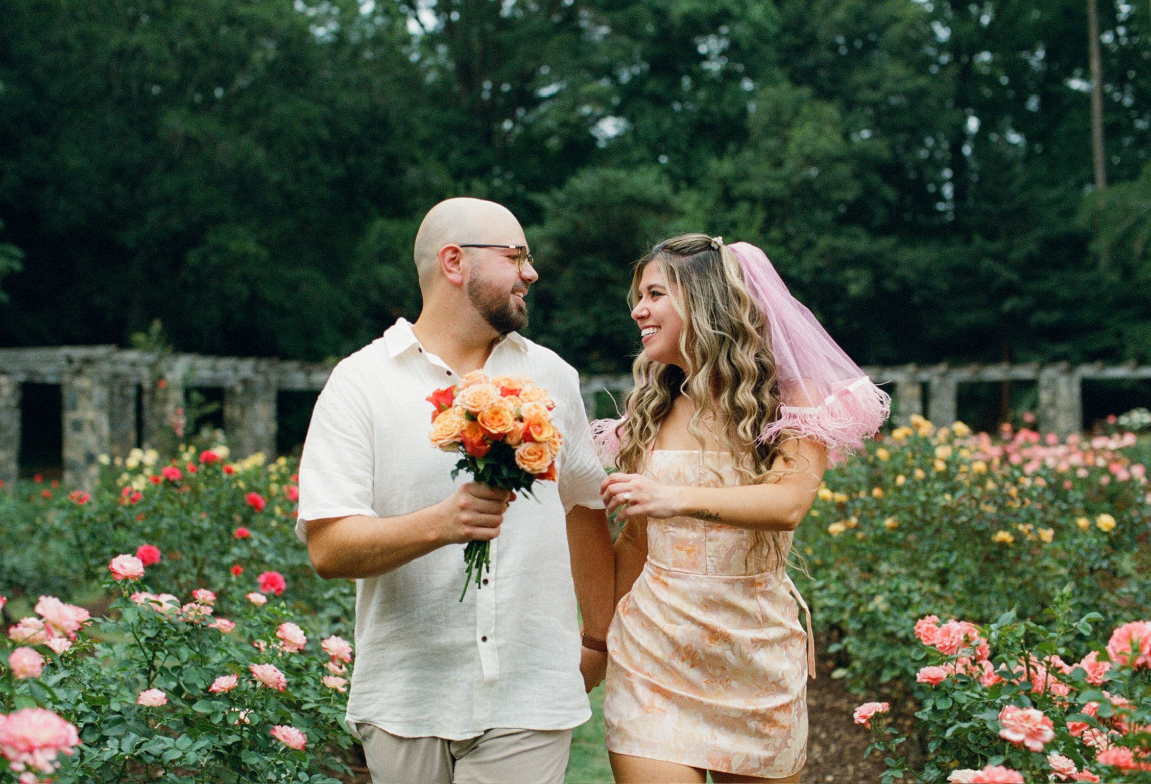 Ian carrying an orange bouquet as Nicole, in her pink veil, smiles up at him among the rose beds.