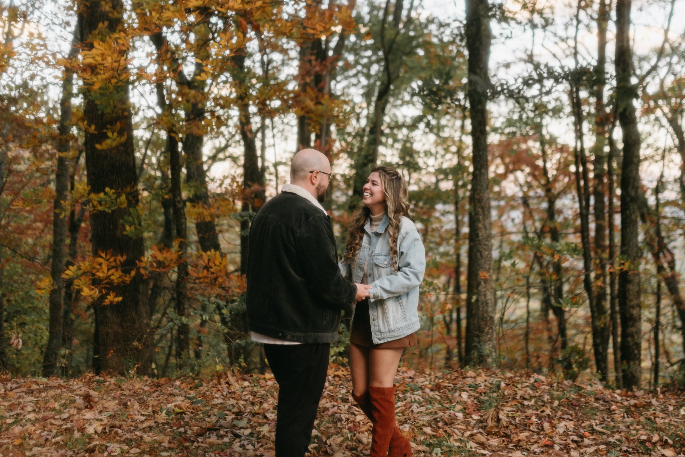 Nicole and Ian facing each other in the autumn woods, leaves underfoot.