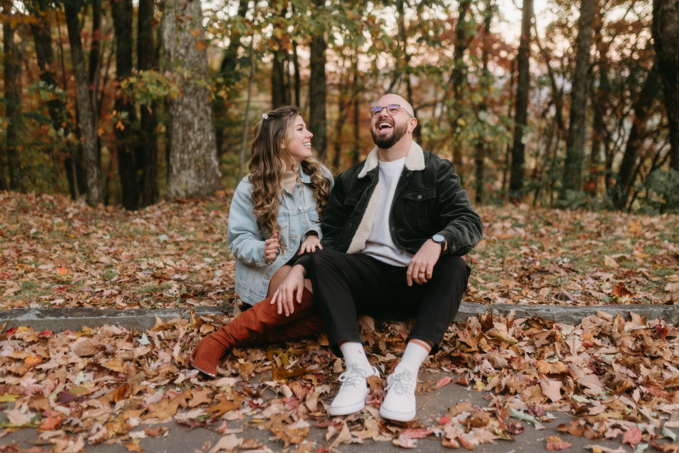 Nicole and Ian sitting together on a low stone edge among the leaves, Ian laughing.