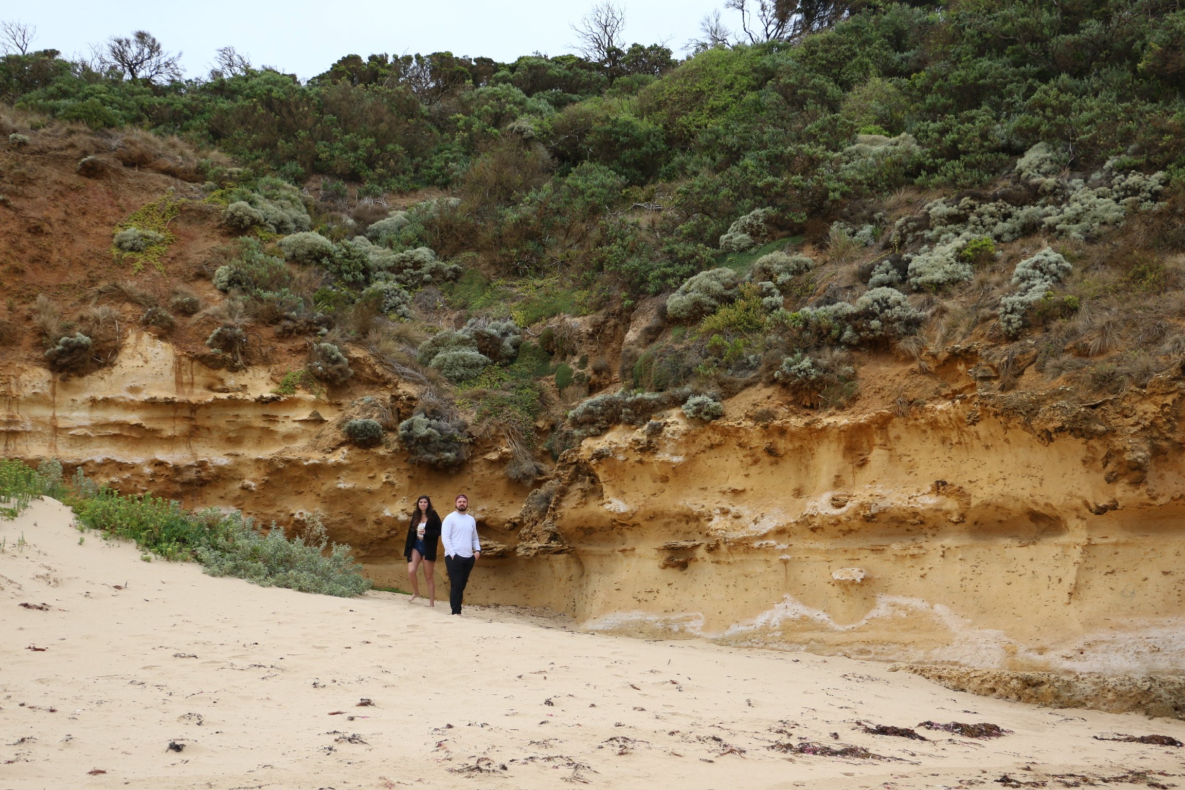 Two small figures walking a sandy cove beneath a sandstone cliff topped with scrub.