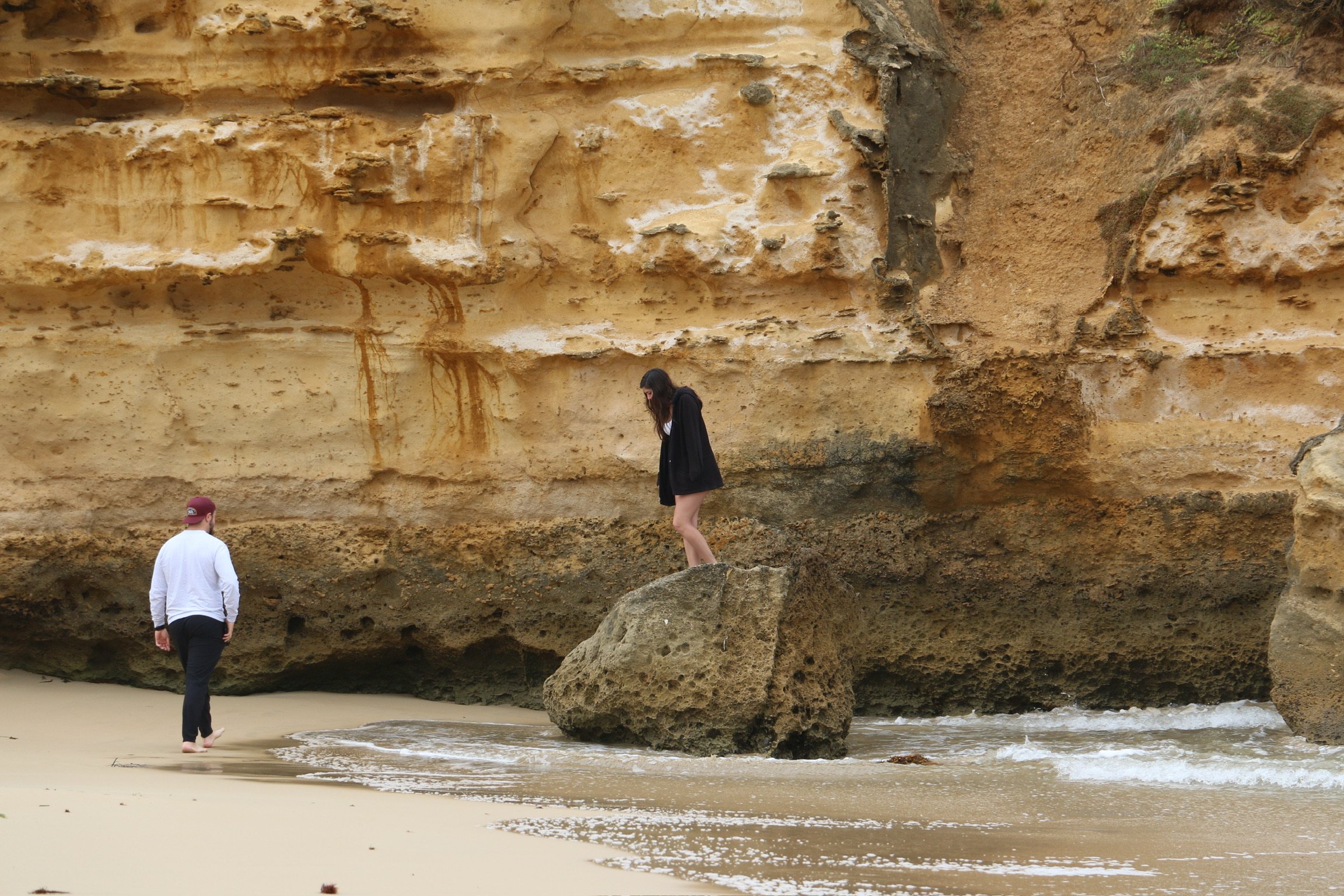 Two people exploring the foot of a sandstone cliff, one climbing onto a boulder at the water’s edge.