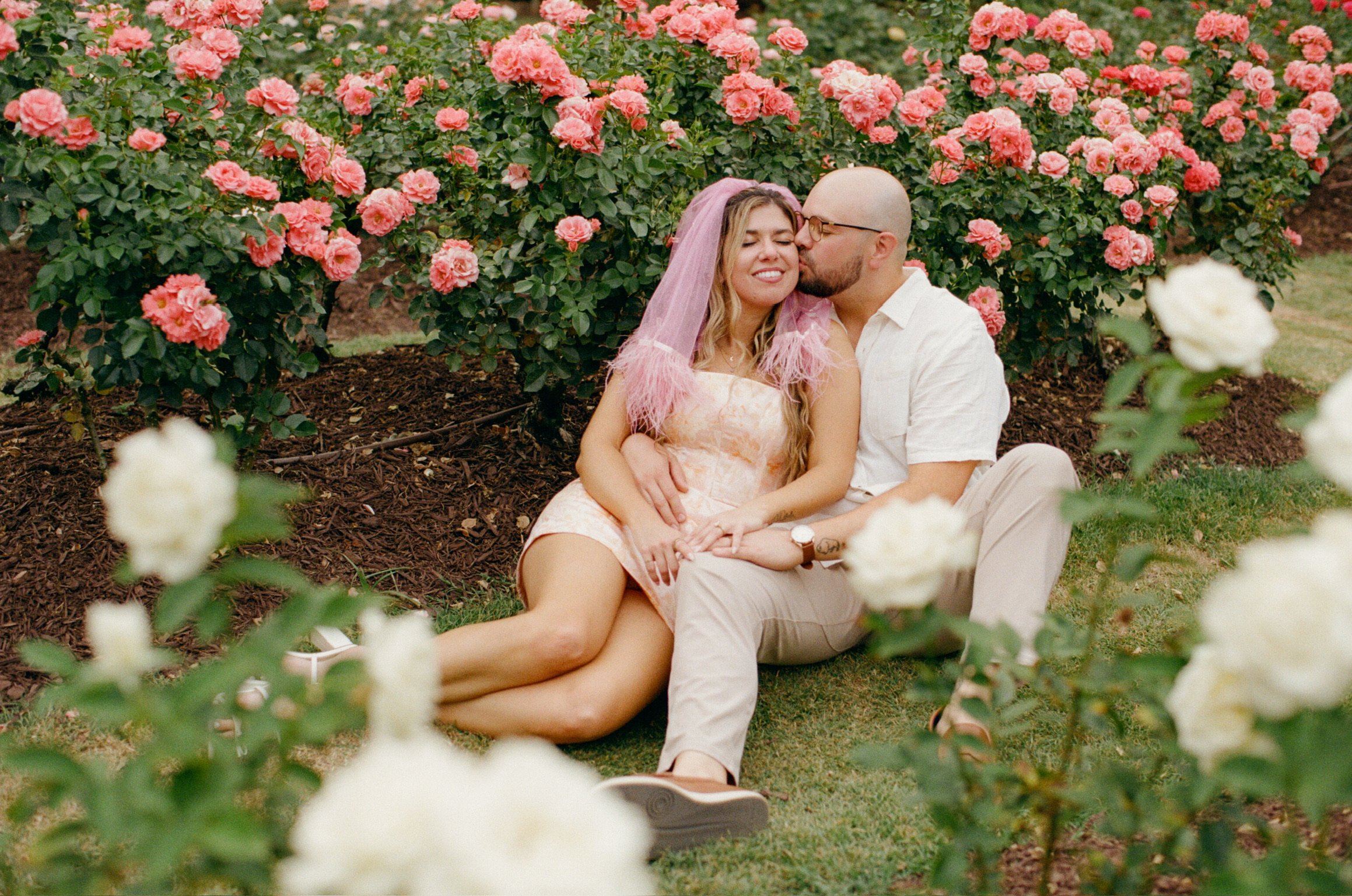 Ian kissing Nicole’s temple as they sit on the grass among pink and white roses.