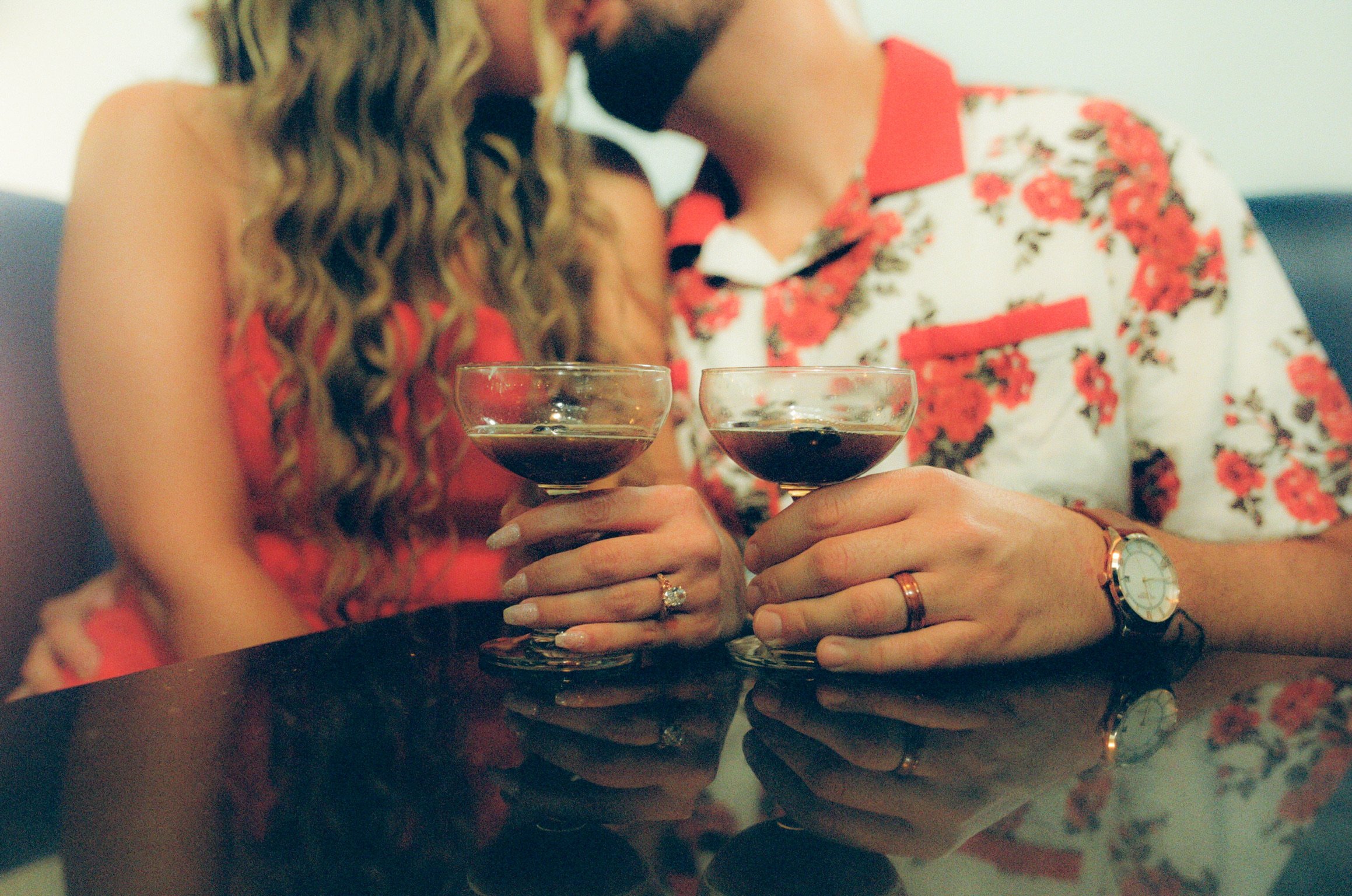 Close up: their two glasses of red wine side by side on the bar, the couple kissing out of focus behind.