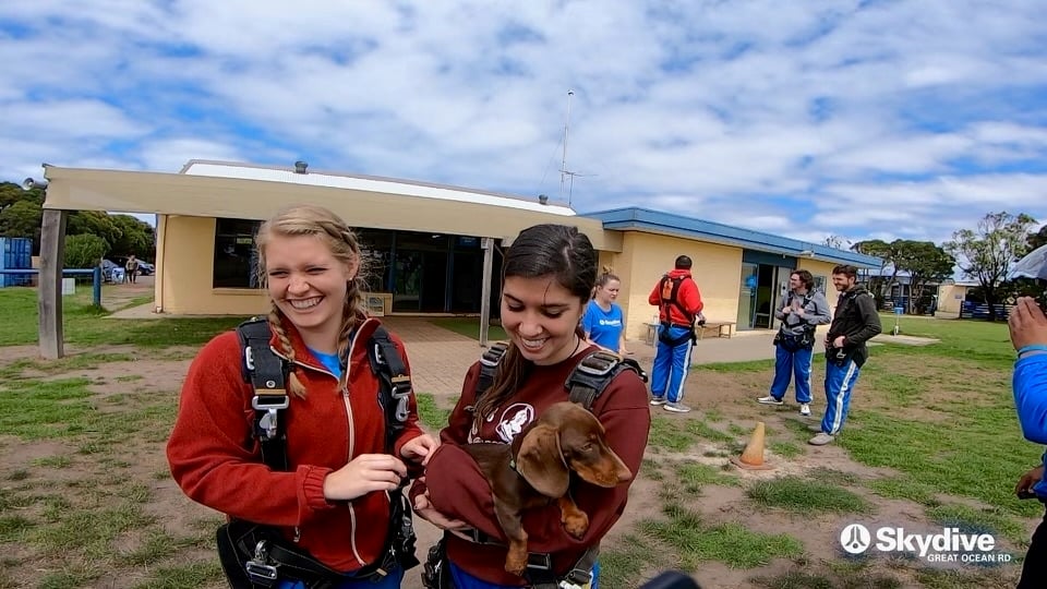 Two women in skydiving harnesses at the landing field, one holding a small brown dachshund.
